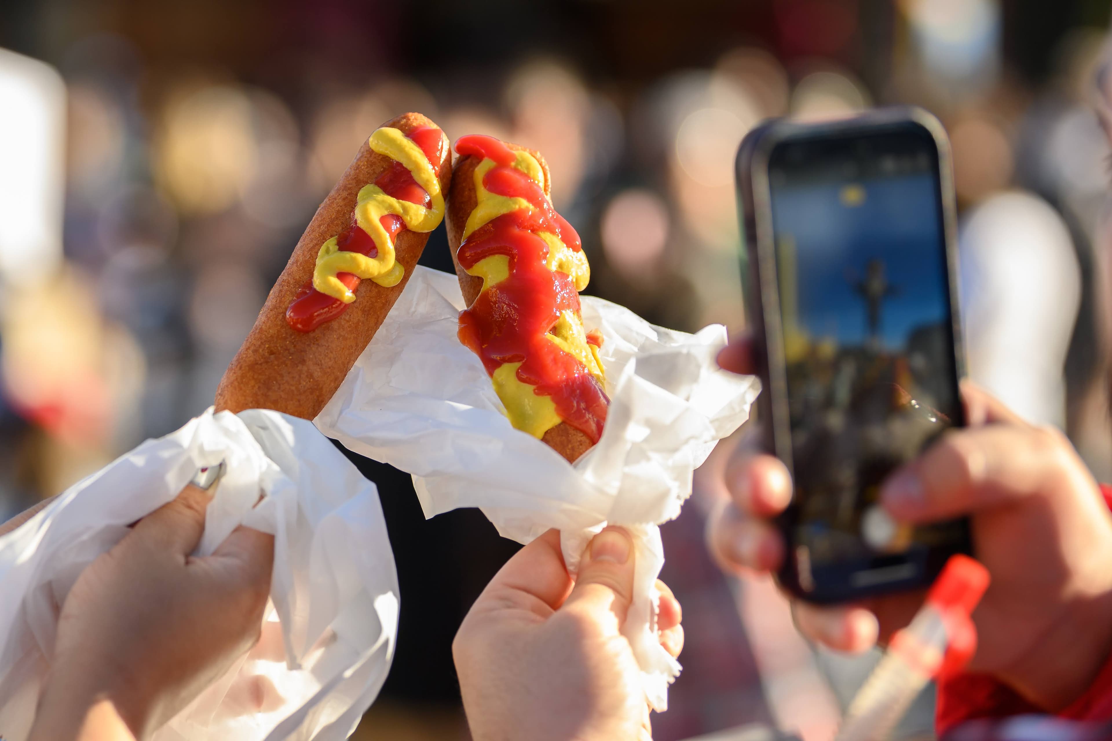 File photo: Fairgoers hold two corny dogs topped with mustard and ketchup; another person photographs them.